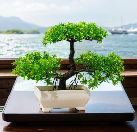 An artificial bonsai tree in a white pot placed on a black surface with a body of water and hills in the background.